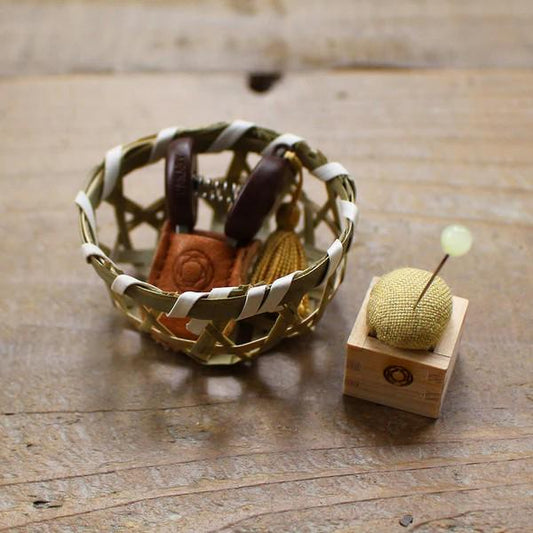 A woven basket containing various small items, such as a tassel, a keychain, and a small wooden box with a needle and thread, sits on a wooden surface.