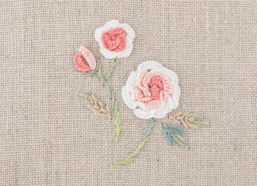 a close-up view of a beige fabric with three crocheted flowers in shades of pink and white, along with some green leaves.