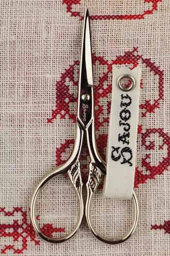 A pair of silver scissors with a white tag attached to them, placed on a red and white embroidered fabric.