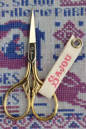 A pair of gold-colored scissors with a white tag attached, resting on a colorful embroidered background.