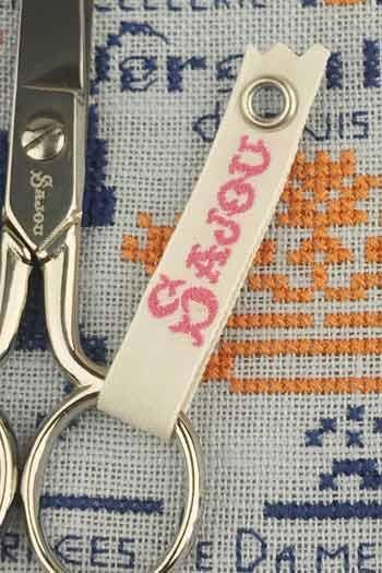 A pair of scissors with a white leather keychain attached to it, featuring a pink text logo.