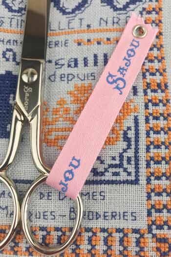 A pair of scissors with a pink tag attached to them, resting on a cross-stitch pattern.