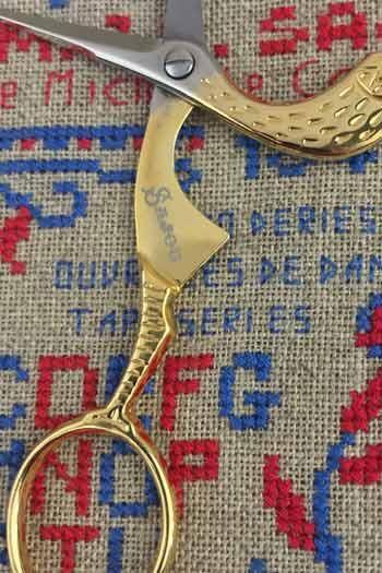A pair of gold-colored scissors with a textured handle is shown on a textured fabric background.