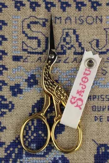 A pair of gold-colored scissors with a bird-shaped handle is placed on a cross-stitch patterned fabric.