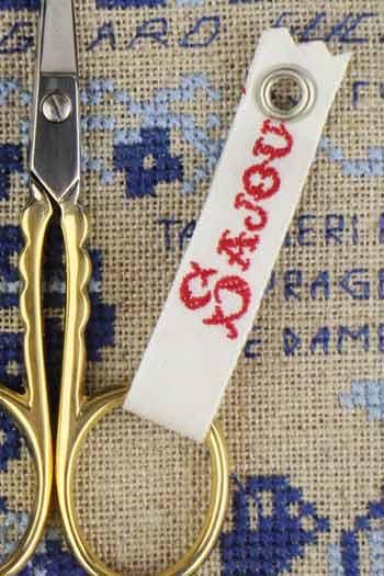 A pair of gold scissors with a white tag featuring red text is placed on a beige fabric with blue and white embroidered patterns.