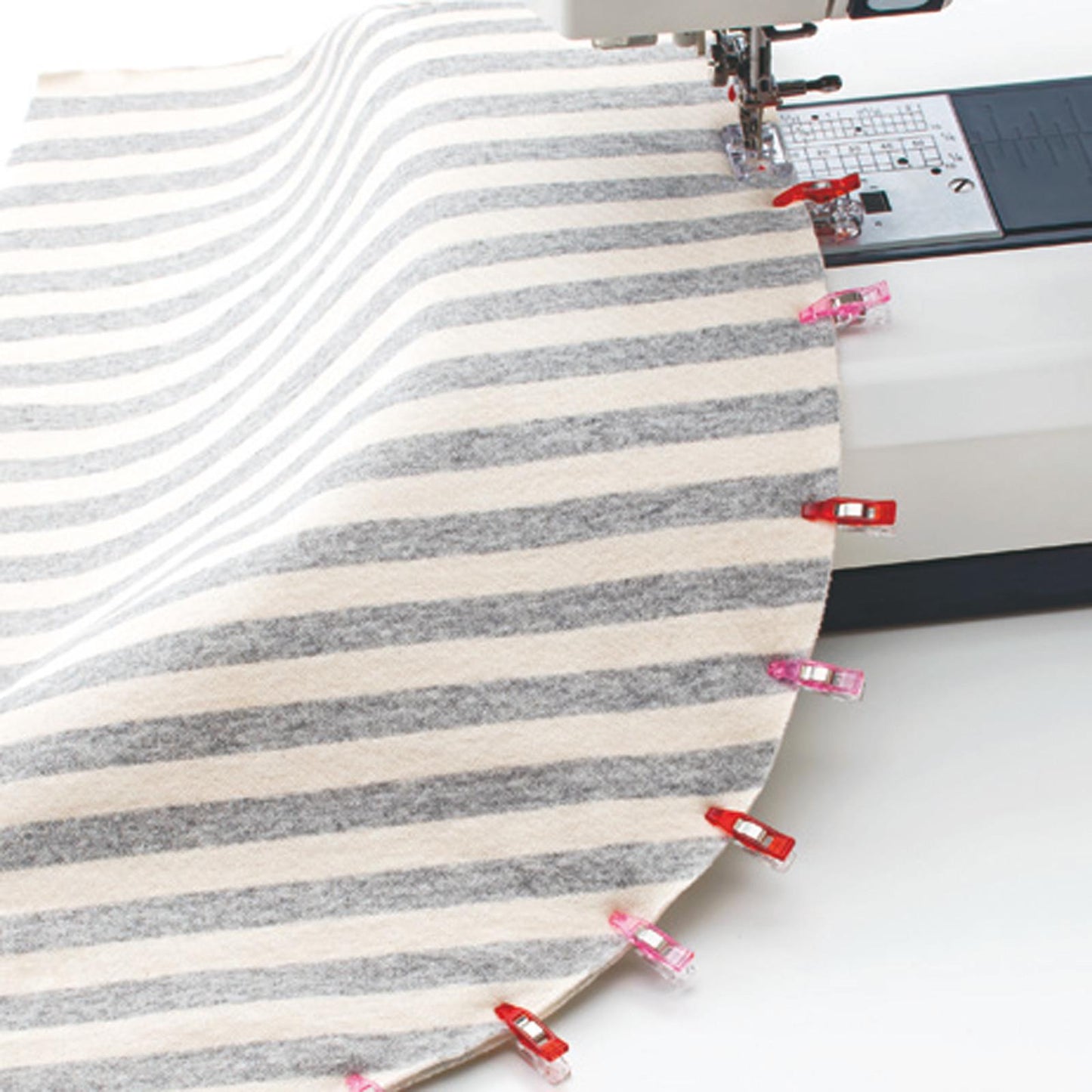 A sewing machine is in use, with a gray and white striped fabric being sewn onto a round object. The fabric is secured with red clips, and the sewing machine is positioned on the right side of the image.