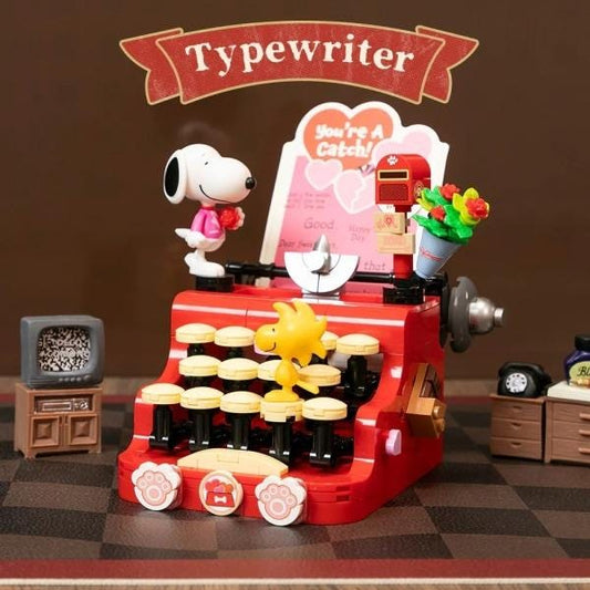 A red toy typewriter with a cartoon character, likely a Peanuts character, sitting on top of it. The typewriter has a small television and a toy mailbox on it, and there are other small toy items around the typewriter.