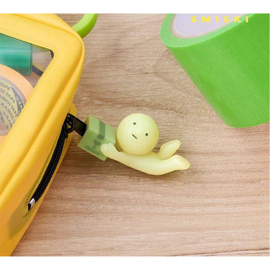 A small, yellow figurine is sitting on a wooden surface next to a green object, possibly a piece of tape.
