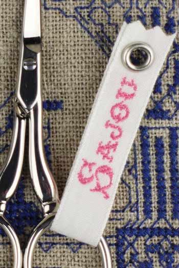 A pair of silver scissors with a white tag featuring the word "Sofa" printed on it, placed on a beige surface with blue embroidered patterns.