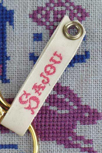 A close-up image of a beige keychain with a red text label attached to it, resting on a fabric with a purple and blue pattern.