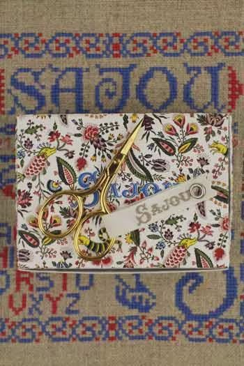 A pair of gold scissors rests on a white fabric with a floral pattern, placed on a beige background with blue and red embroidered designs.
