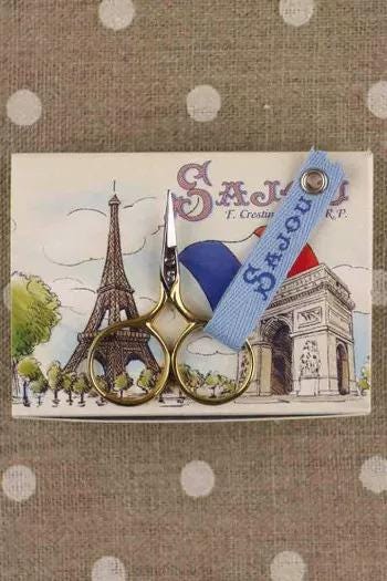 A pair of gold scissors with a blue handle and a label that says "Sagou" is placed on a card featuring an illustration of the Eiffel Tower.