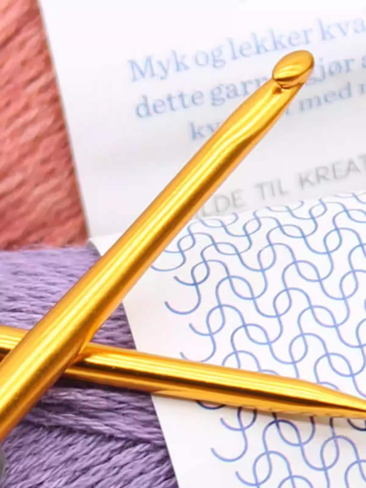 a close-up view of a gold crochet hook and a ball of purple yarn, with a partially visible piece of paper with blue and white text in the background.