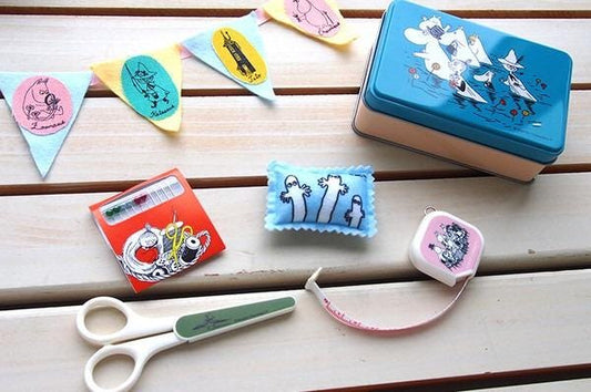 various craft supplies and decorative items, including a pair of scissors, a small blue and white pillow, a blue and white tin, and a pink and white measuring tape.