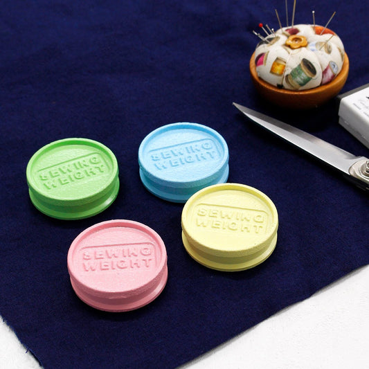 four colorful sewing weight containers, each with a different color, placed on a blue surface alongside a pair of scissors and a needle and thread.