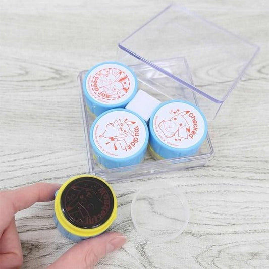 A hand is holding a small, round object with a yellow and blue color scheme, which appears to be a stamp or a similar item. The object is placed next to a clear plastic container with three blue and red items inside, which are also placed on a wooden surface.