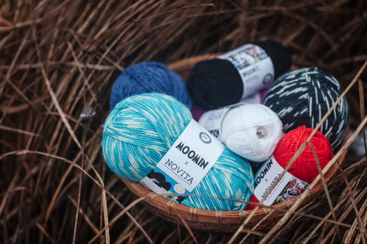 A basket filled with various balls of yarn, including blue, black, white, and red yarn, resting on a bed of straw or grass.