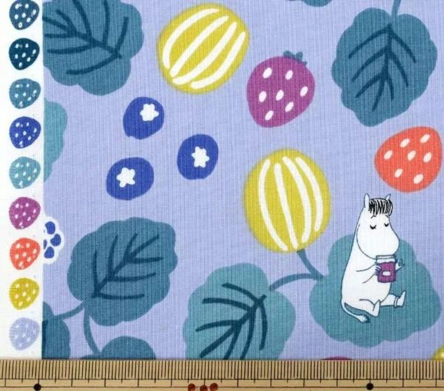 a fabric with a playful pattern of fruits and leaves, including strawberries, blueberries, and a small white character. The fabric is displayed on a wooden ruler, which is placed on a wooden surface.