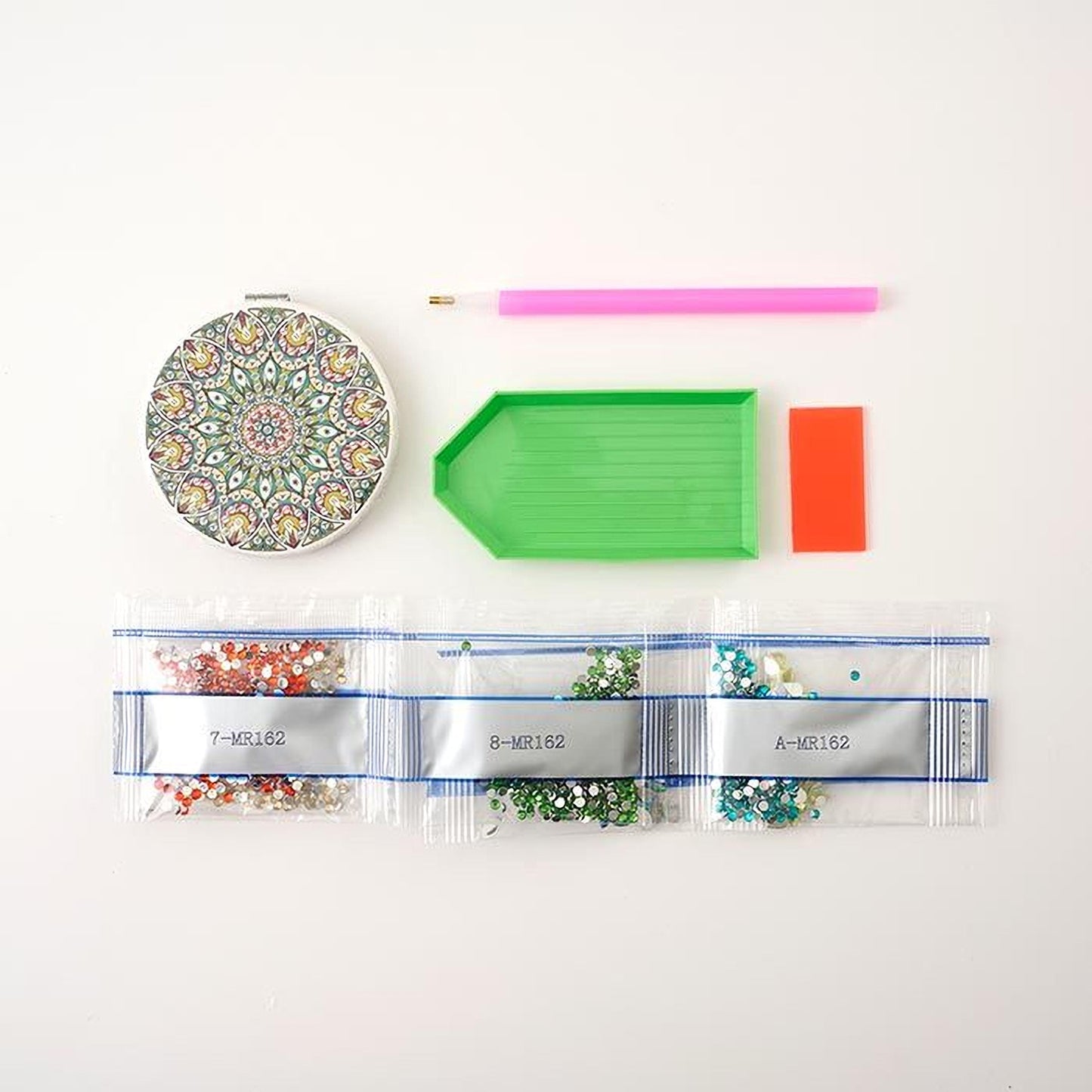 various craft supplies, including a round mirror with a colorful design, a pink pen, a green plastic tray, and three clear plastic bags filled with colorful beads or sequins.