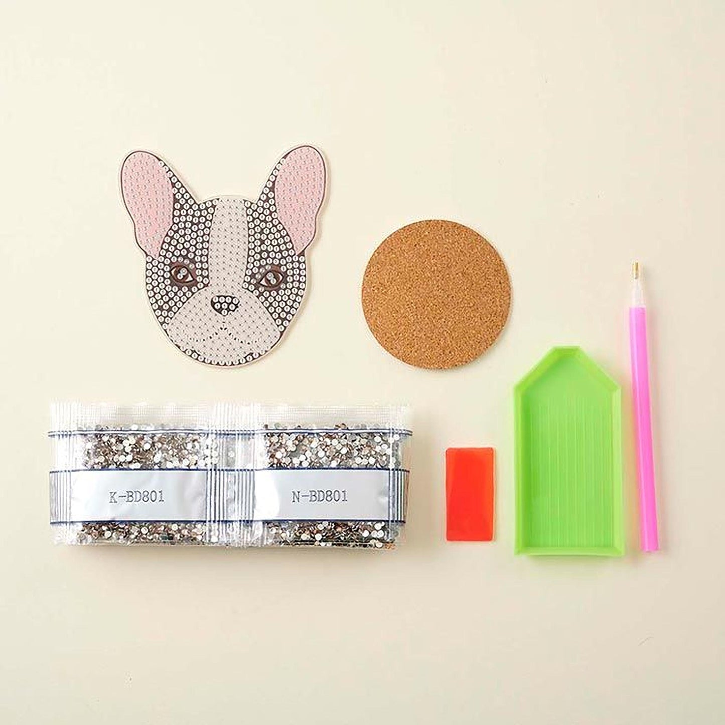 various craft supplies, including a glittery dog coaster, a cork coaster, a pen, a diamond tool, and a package of glitter beads.