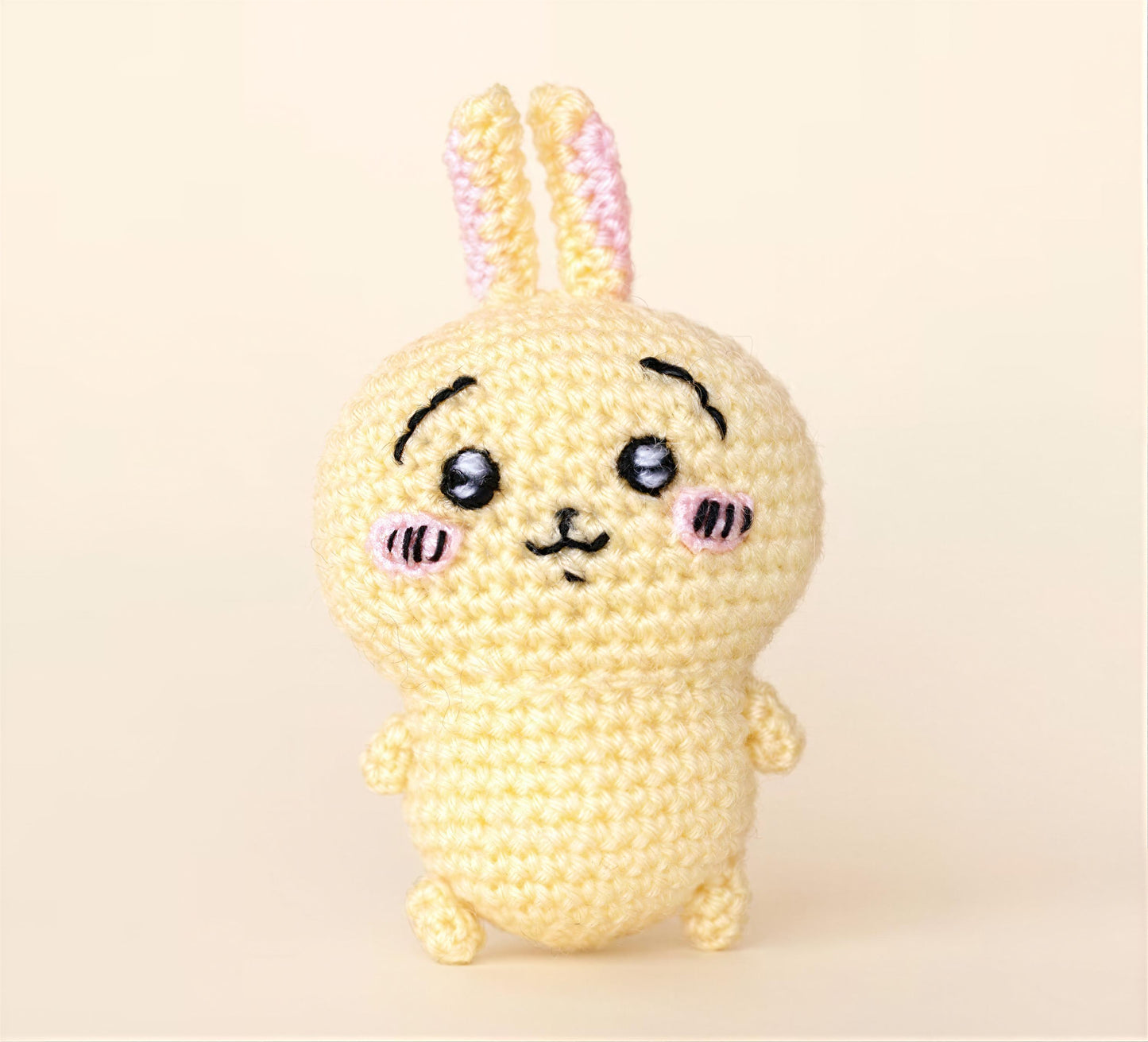 A crocheted toy rabbit with a cute face and pink ears is sitting on a plain background.