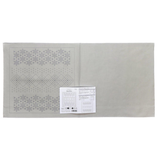 The image is a gray fabric with a repeating pattern of snowflakes on it. There is a white paper with text and a barcode attached to the fabric.