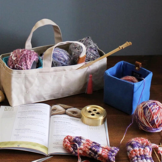 A white tote bag filled with yarn and knitting needles sits on a wooden surface, accompanied by a book and various knitting supplies.