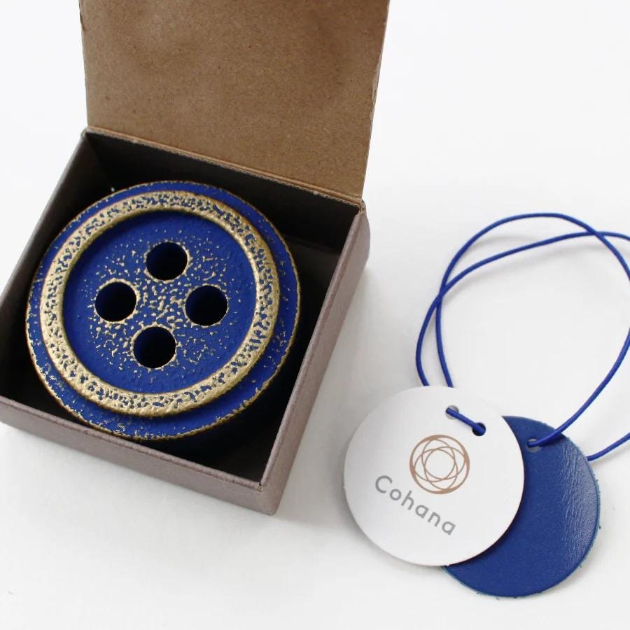 a blue button with gold accents placed inside a brown cardboard box, accompanied by a white tag with the word "Cohana" printed on it.