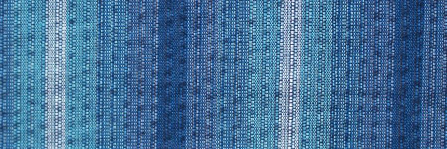a close-up view of a blue and white striped fabric, with the fabric appearing to be woven or knitted. The fabric has a textured and patterned appearance, with vertical stripes in shades of blue and white. The image does not contain any text or other discernible objects or actions.