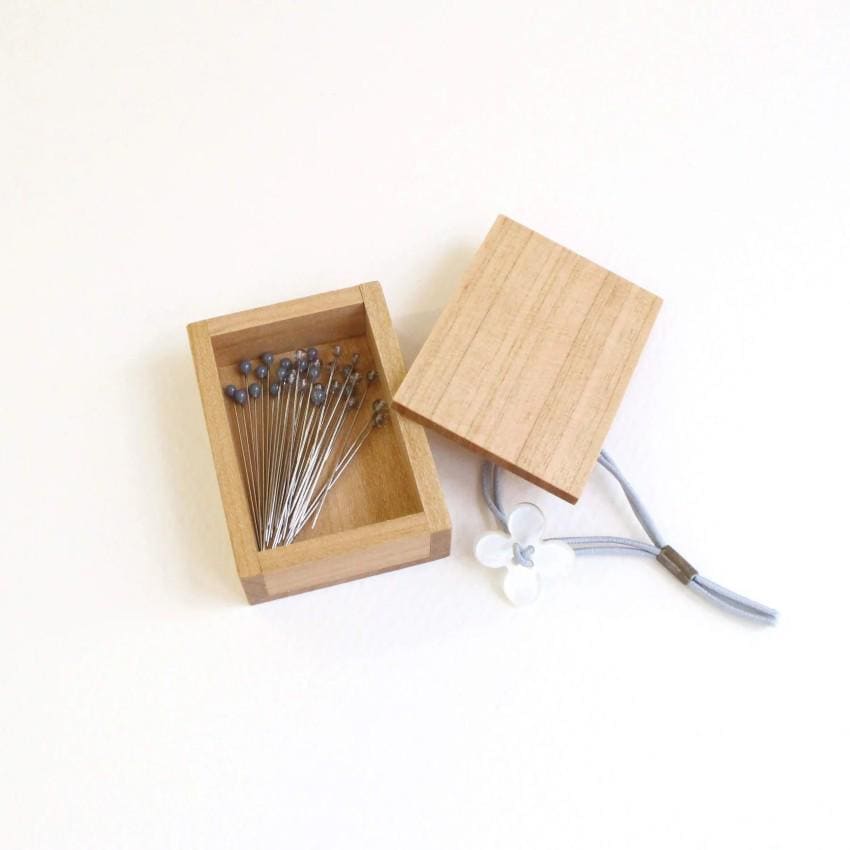 A wooden box containing a collection of small pins, with a clear plastic bag attached to the lid.