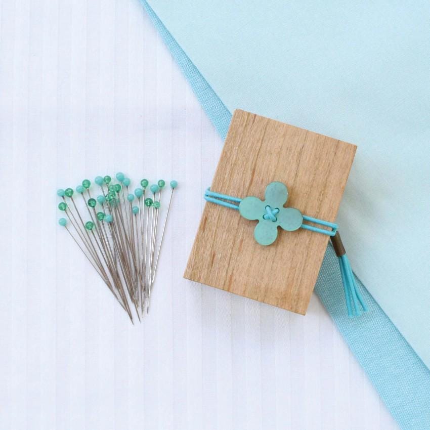 a wooden block with a blue flower-shaped button on it, surrounded by a collection of green push pins and a blue fabric.