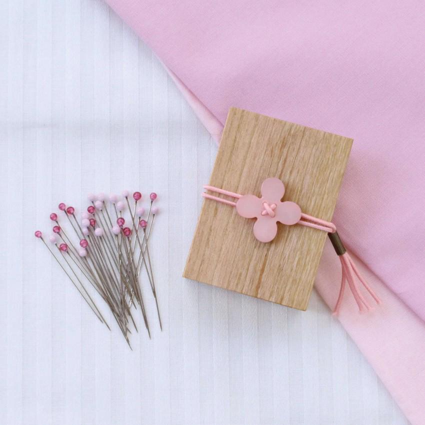 a wooden box with a pink flower-shaped button on it, surrounded by a pile of pins and a pink fabric.