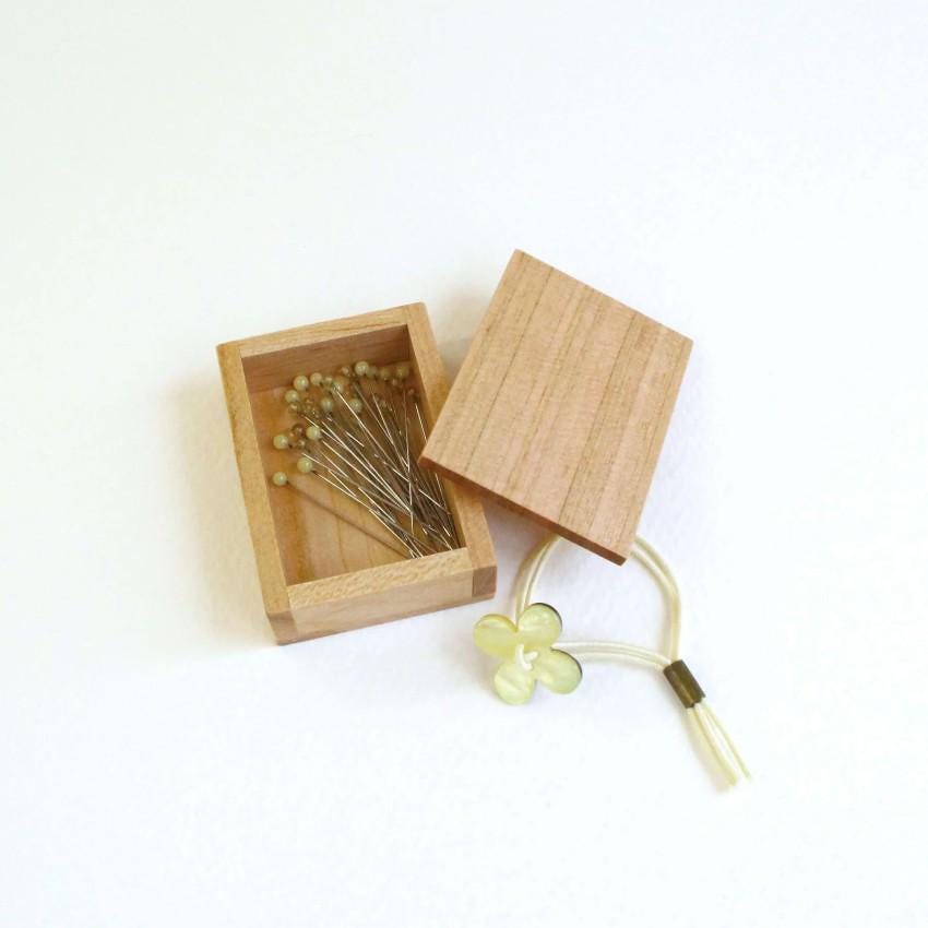 a wooden box with a lid, containing several small pins, and a small yellow flower-shaped object with a white string attached to it.
