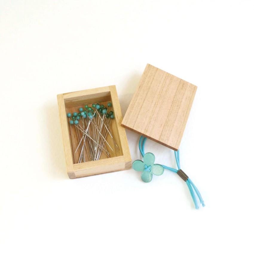 A wooden box containing a collection of blue pins and a small blue flower-shaped charm.
