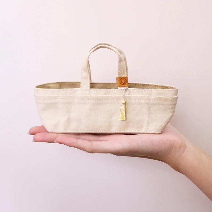 A hand holding a small, beige-colored tote bag with a tassel attached.