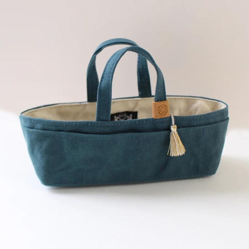 A teal-colored tote bag with a tassel hanging from the handle, sitting on a white surface.