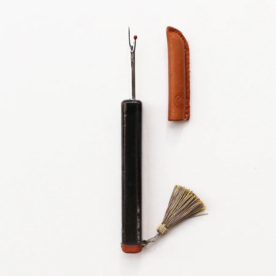 A black sewing needle with a tassel attached, placed next to a brown leather tool case.