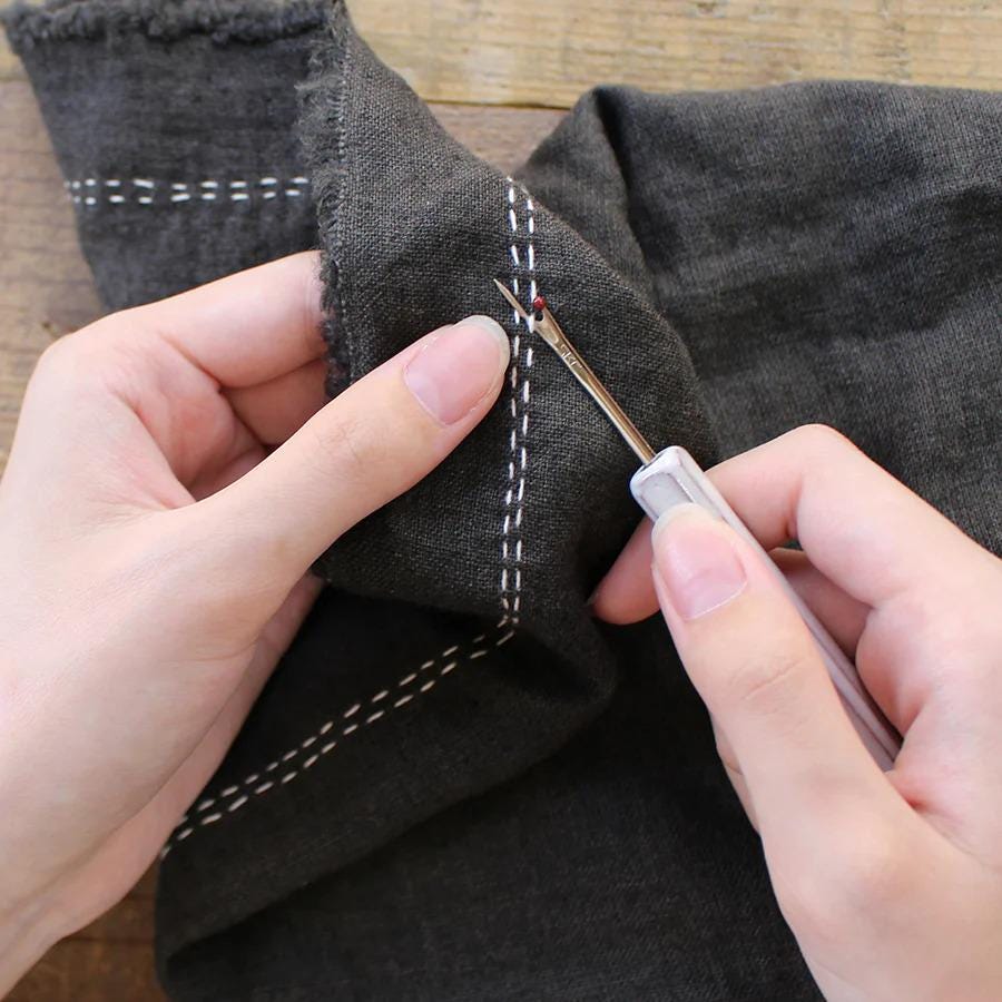 A pair of hands holding a sewing needle and thread, carefully stitching a fabric with white stitching.