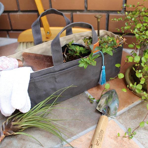 A gray tote bag filled with various gardening tools and plants sits on a tiled surface, with a potted plant nearby.