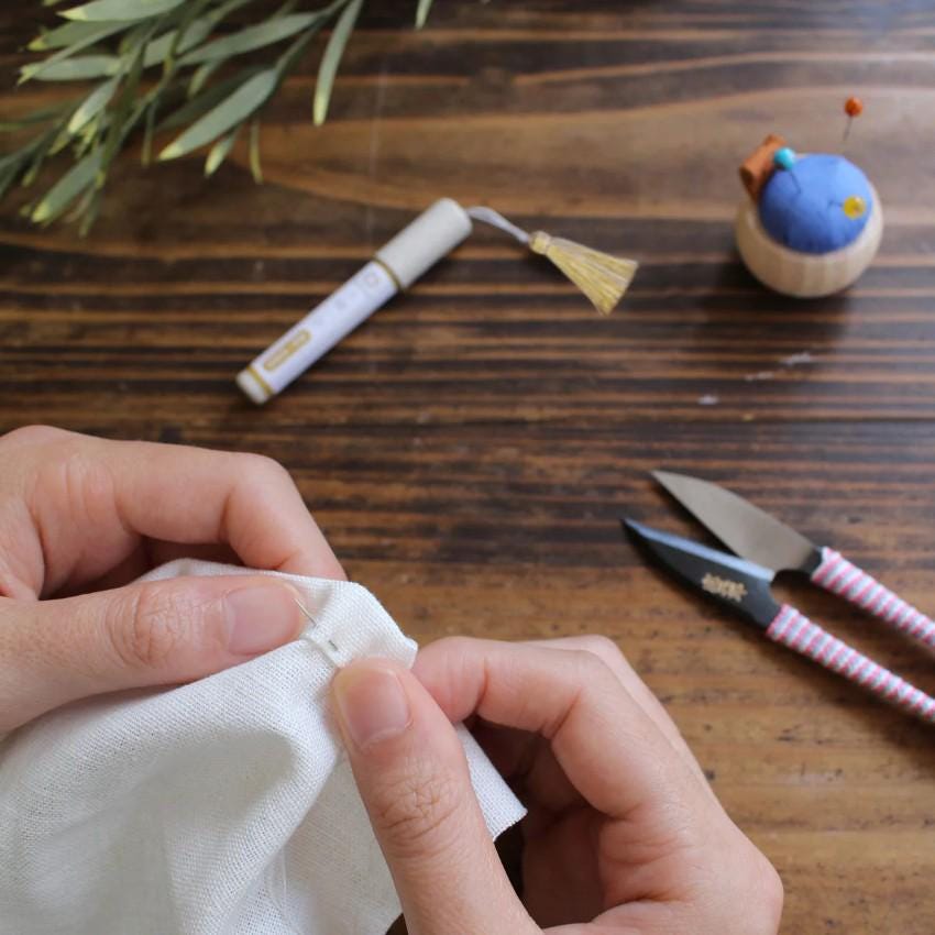 A person's hands are carefully stitching a piece of fabric on a wooden table, with a pair of scissors and a needle and thread nearby.