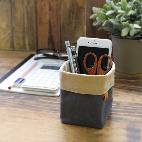 A wooden desk with various office supplies, including a calculator, a pair of glasses, a smartphone, and a pair of scissors, all neatly organized in a small gray and beige container.