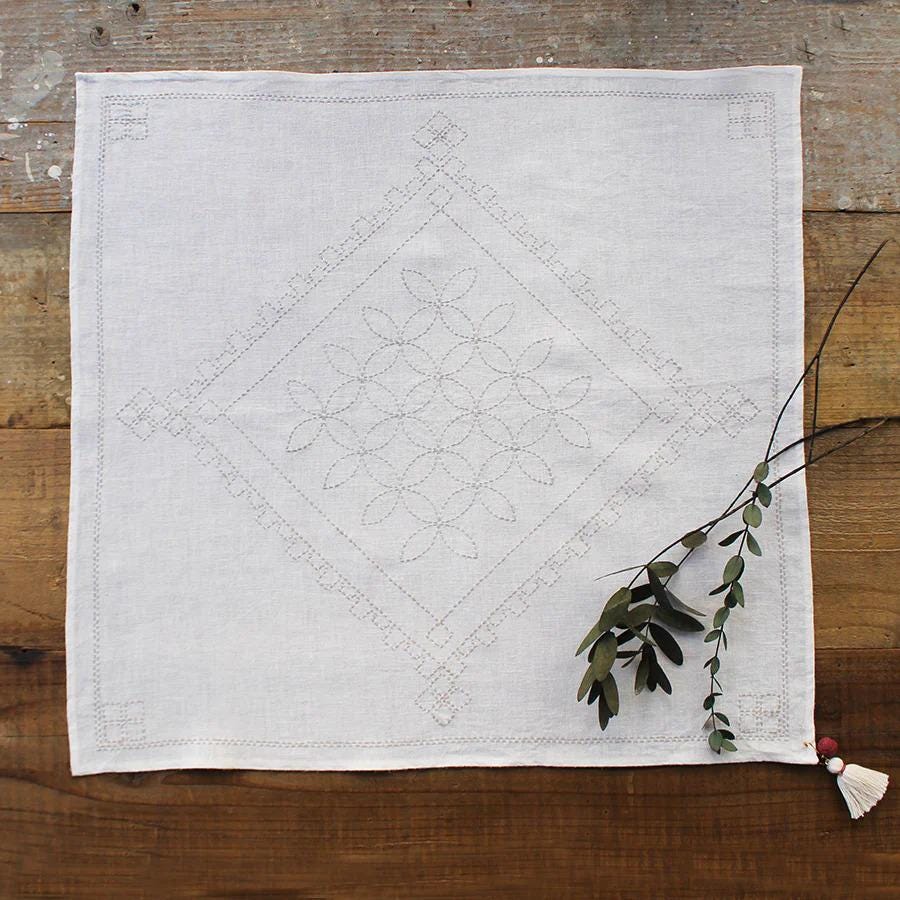 a white square napkin with a decorative floral pattern, placed on a wooden surface alongside a branch with green leaves.