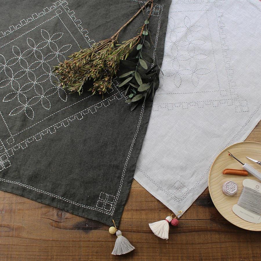 a wooden table with a gray tablecloth, a white tablecloth, and some embroidery supplies, including a needle, thread, and tassels.
