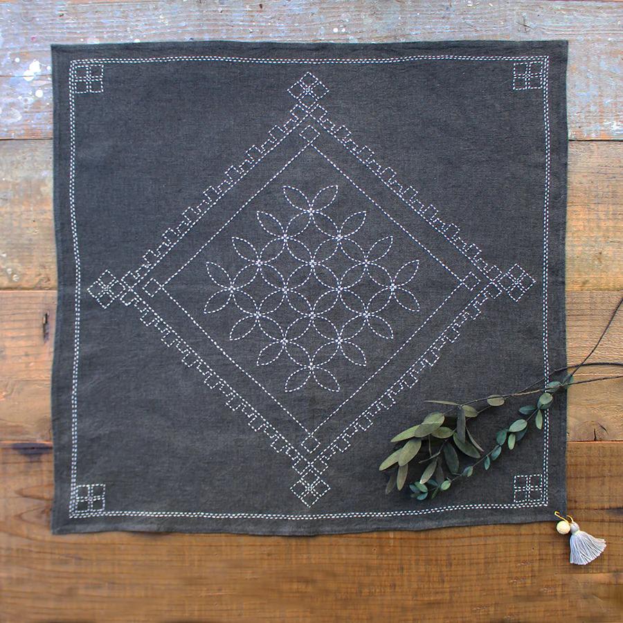 a gray fabric with a white floral design, placed on a wooden surface. Next to the fabric, there is a branch of green leaves.