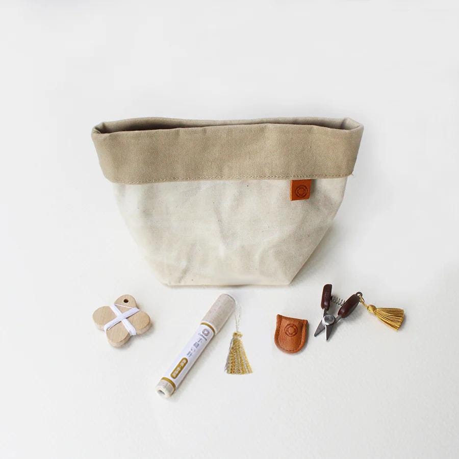 A beige canvas bag with a brown trim, a white marker, a pair of scissors, and some tassels are arranged on a white background.