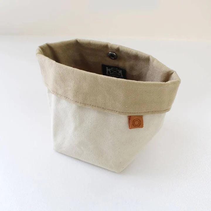 A beige canvas tote bag with a brown flap and a small orange tag attached, sitting on a white surface.