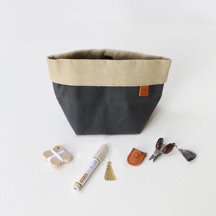 A black and beige tote bag with a leather tag, a white marker, a pair of scissors, and some tassels or trinkets are arranged on a white background.