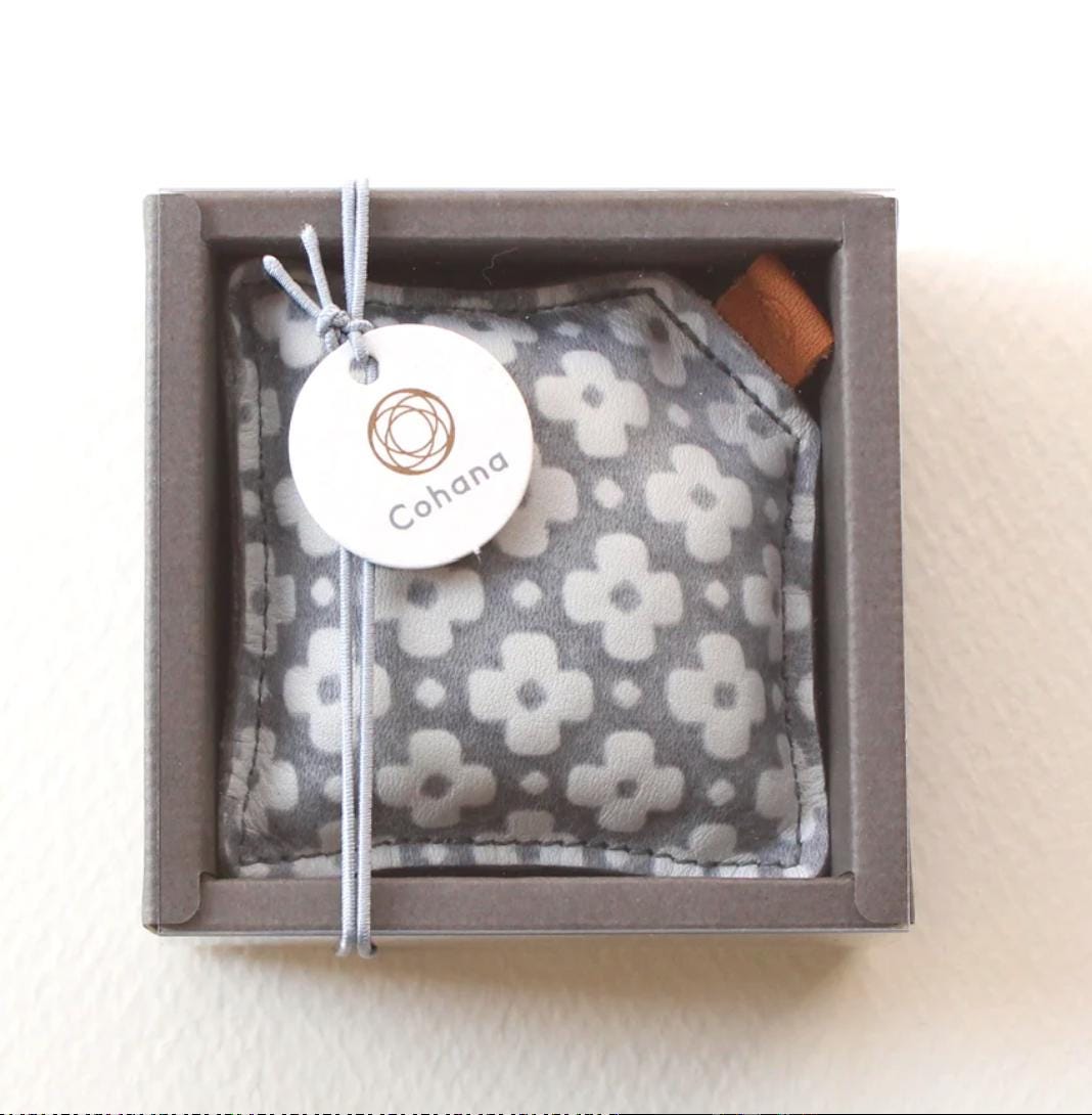 A gray and white patterned fabric item, possibly a cushion or pillow, is displayed in a gray cardboard box with a white circular tag that reads "Cohana".