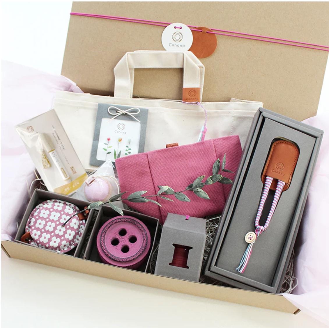 A gift box containing various items, including a white tote bag, a pink bag, a small pink bag, a gray box, a pink button, and a brown leather keychain.
