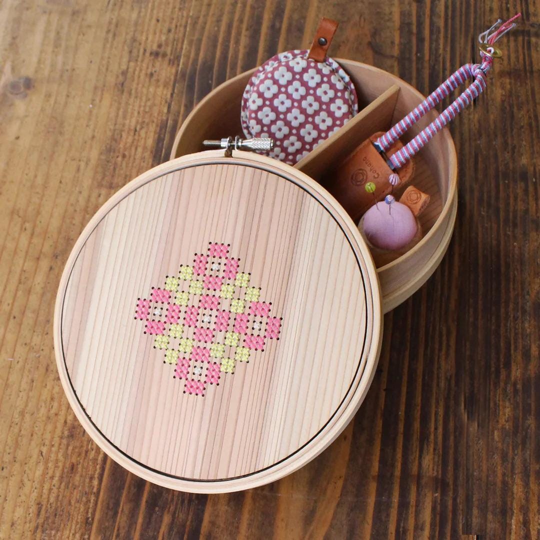 A wooden box with a round lid, containing various small items such as a pink and white flower-patterned pouch, a small pink ball, and a small brown leather object.